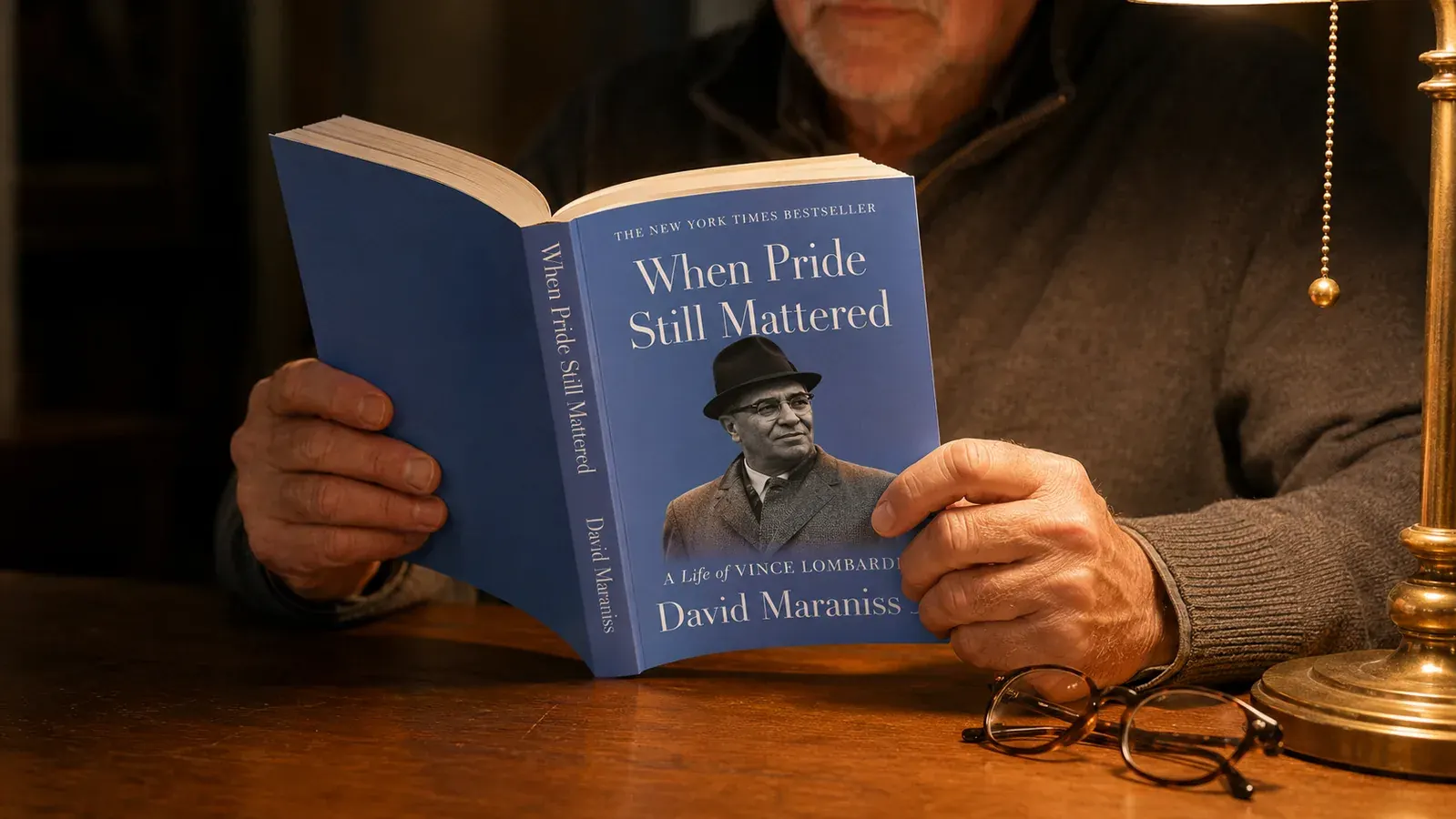 An older reader's hands holding an open thick book on a wooden table under a reading lamp
